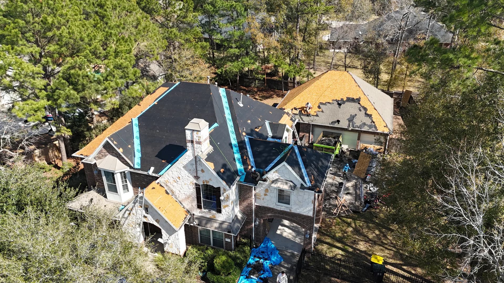 Aerial view of a large two-story home mid-tear-off with exposed decking, underlayment, and crew working