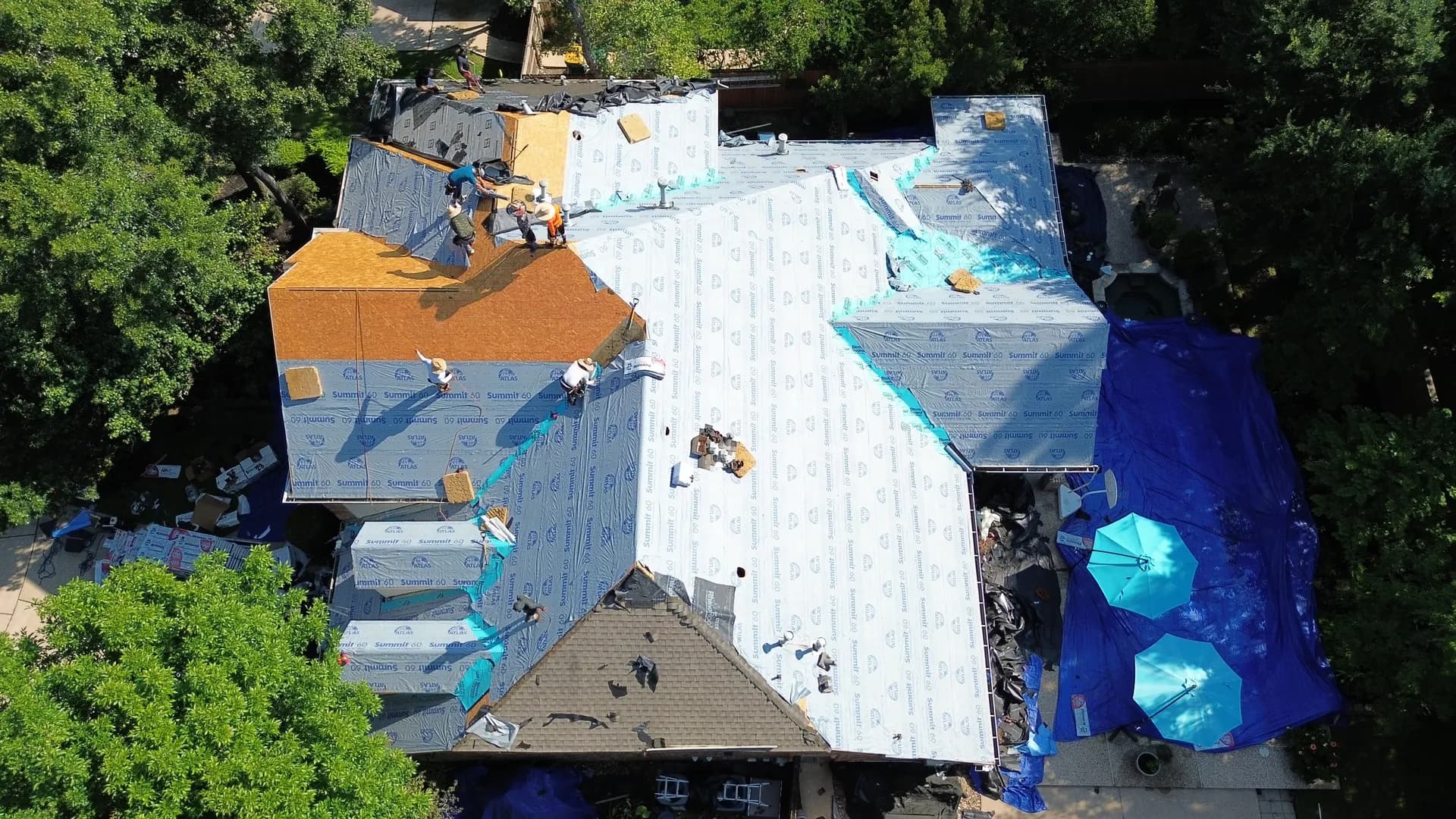 Documentary aerial photo of a roof mid-installation showing synthetic underlayment covering most of the deck, an exposed OSB section, and a crew working, illustrating the sealed roof deck concept central to the FORTIFIED standard.