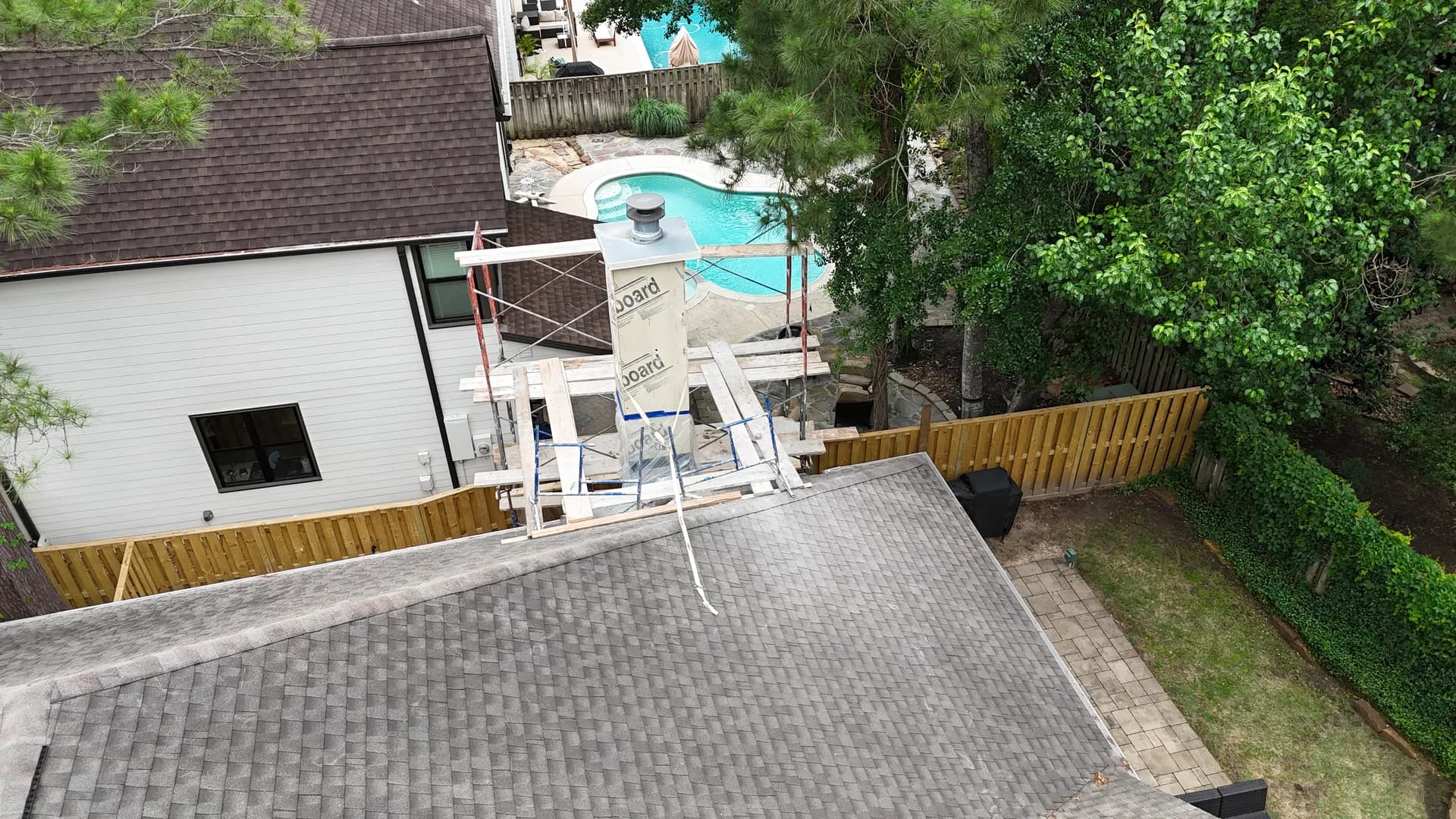 Aerial view of a chimney chase wrapped in insulation board with a new metal cap fitted across the top, scaffolding staged around it on the shingle roof below.