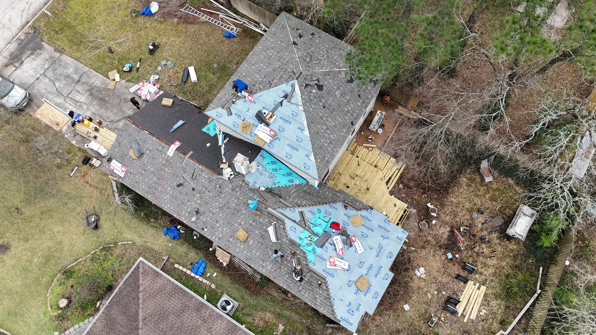 Top-down aerial view of a residential roof mid tear-off, old shingles stripped away, sections of synthetic underlayment already laid down, and a crew working across the roof planes.