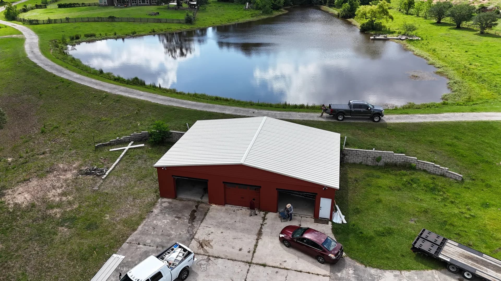 Aerial view of a completed white standing seam metal roof on a red barn beside a pond on acreage