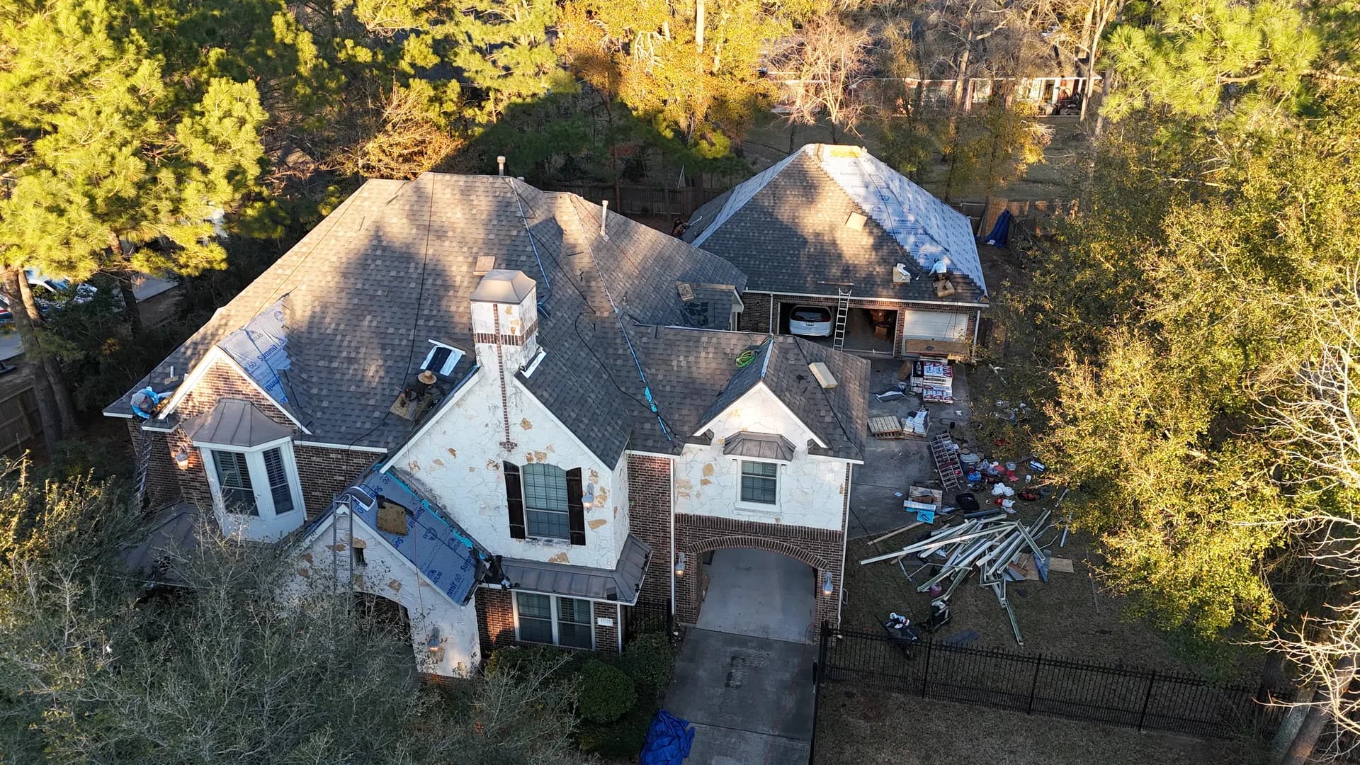 Aerial view of a large brick and stone home mid-assessment, with the roof partially stripped down to underlayment, a cracked chimney tower visible, and roofing materials staged in the driveway, capturing the moment a homeowner has to weigh a repair against a full roof replacement.