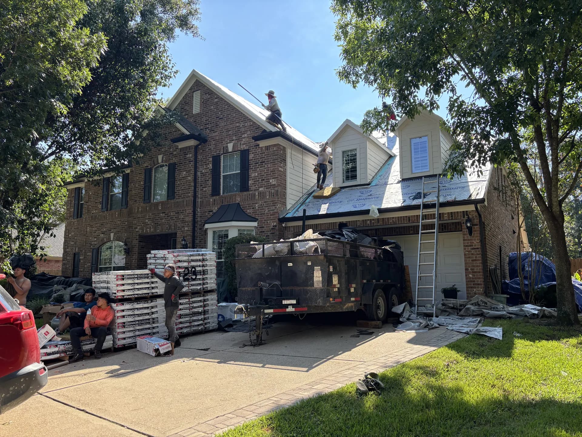 ROYH Build Co crew members working a brick home's roof during a tear-off and reroof, with a debris trailer and shingle bundles staged in the driveway