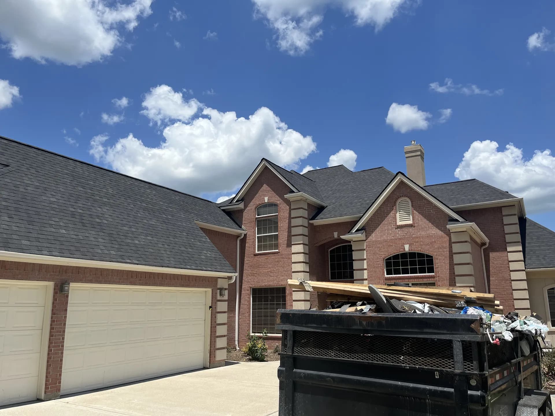 Brick two-story home with a newly shingled roof and a driveway dump trailer full of tear-off debris