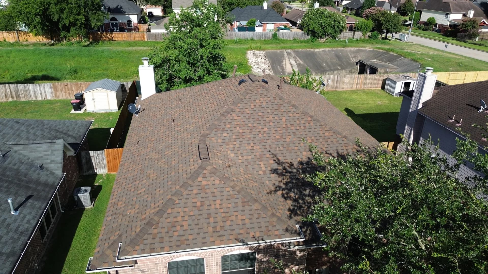 Aerial drone view of a composition shingle roof showing a chimney, roof vents, and a backyard shed