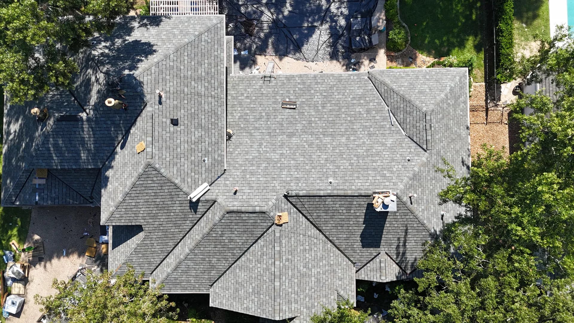 Aerial view of a finished gray shingle roof with crew members visible on the roof and the corner of a pool in the yard below