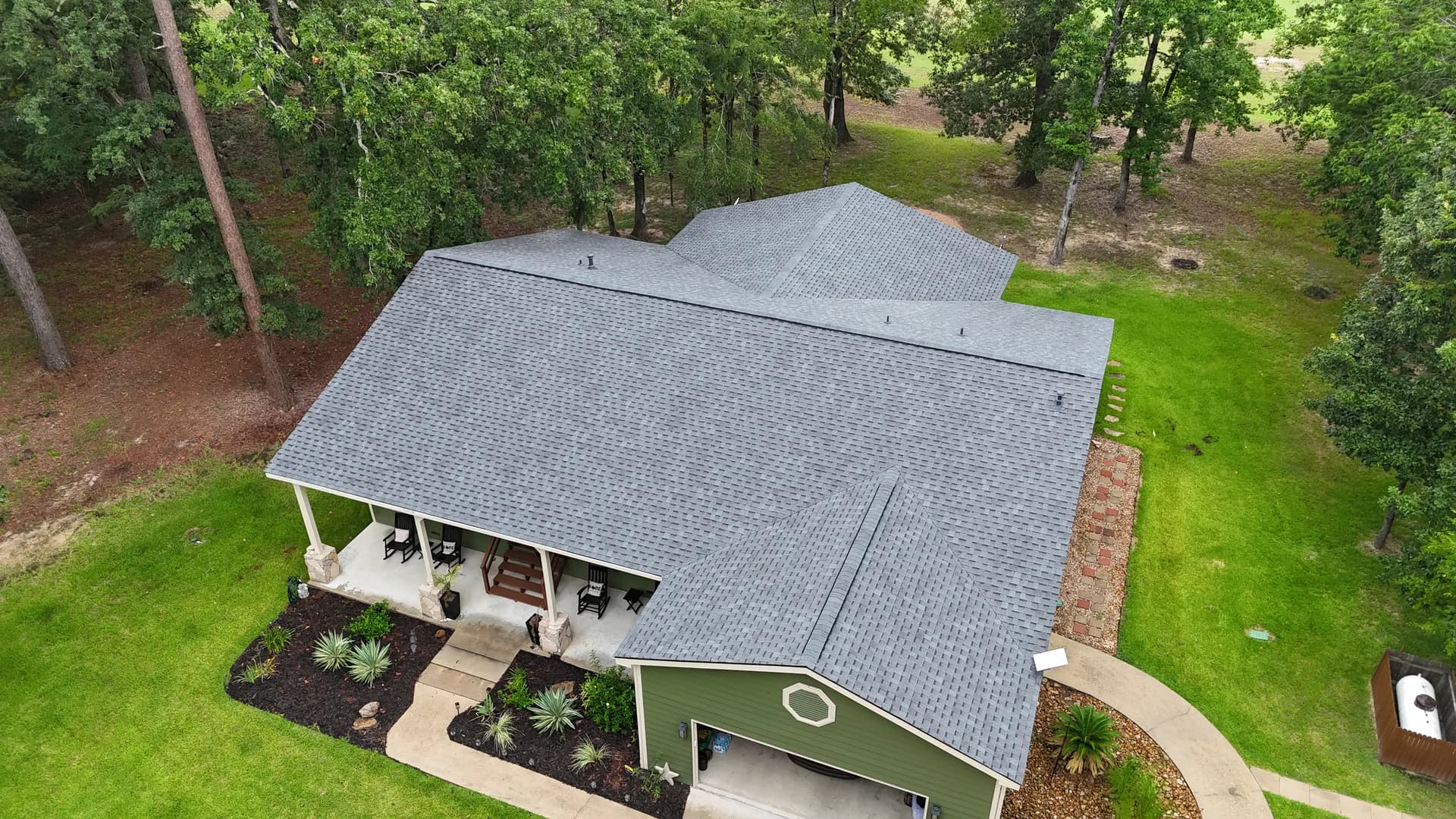 Aerial drone view of a green house with a complex gabled gray shingle roof over a covered front porch and landscaped beds