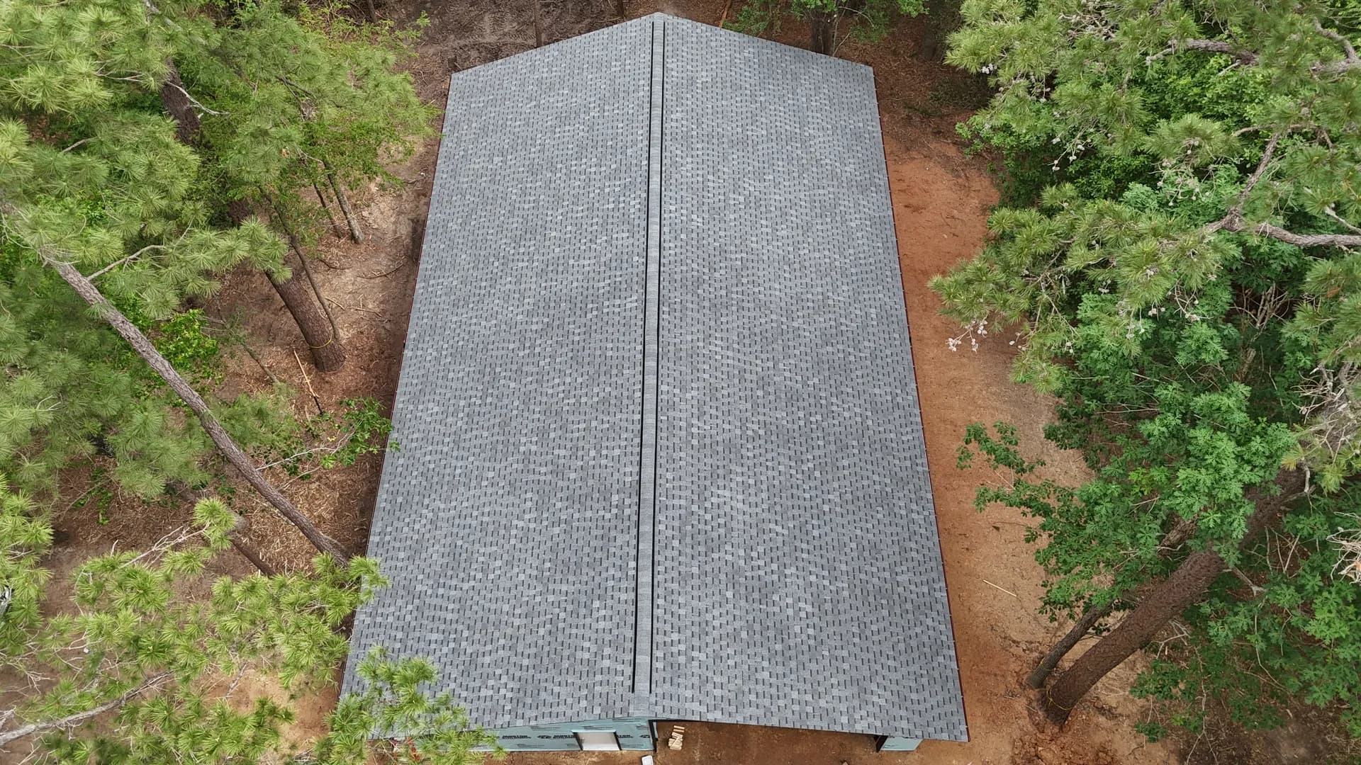 Straight-down aerial of a simple symmetrical gable roof in dark gray architectural shingles flanked by tall pine trees