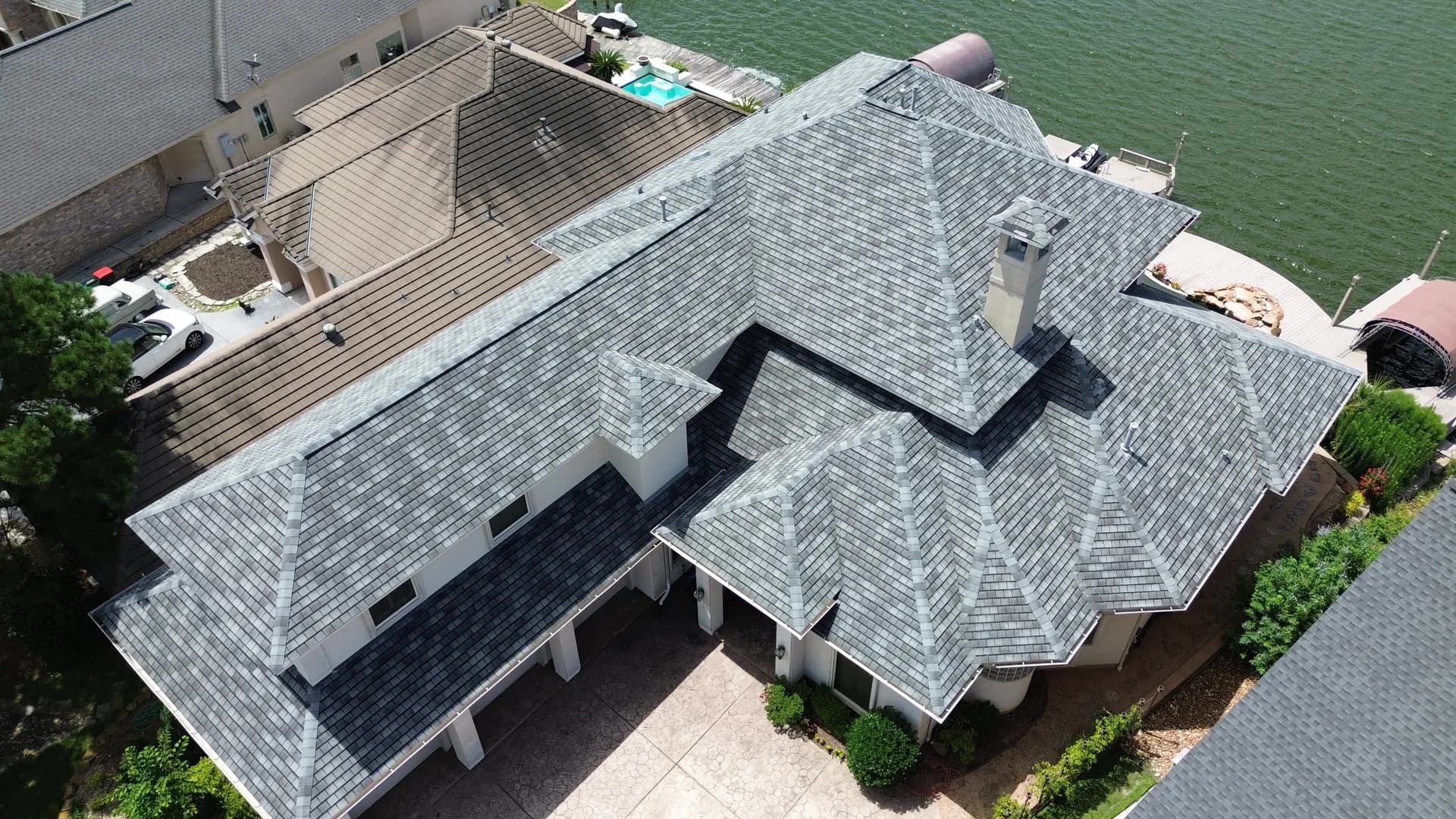 Aerial drone view of a luxury lakefront home with a gray shingle roof, swimming pool, and private boat dock