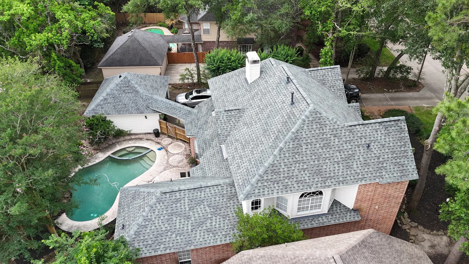 Angled aerial photograph of a residential brick home with a visibly weathered, aged gray asphalt shingle roof showing wear and granule thinning, backyard pool in frame, natural daylight, real photograph not a render.