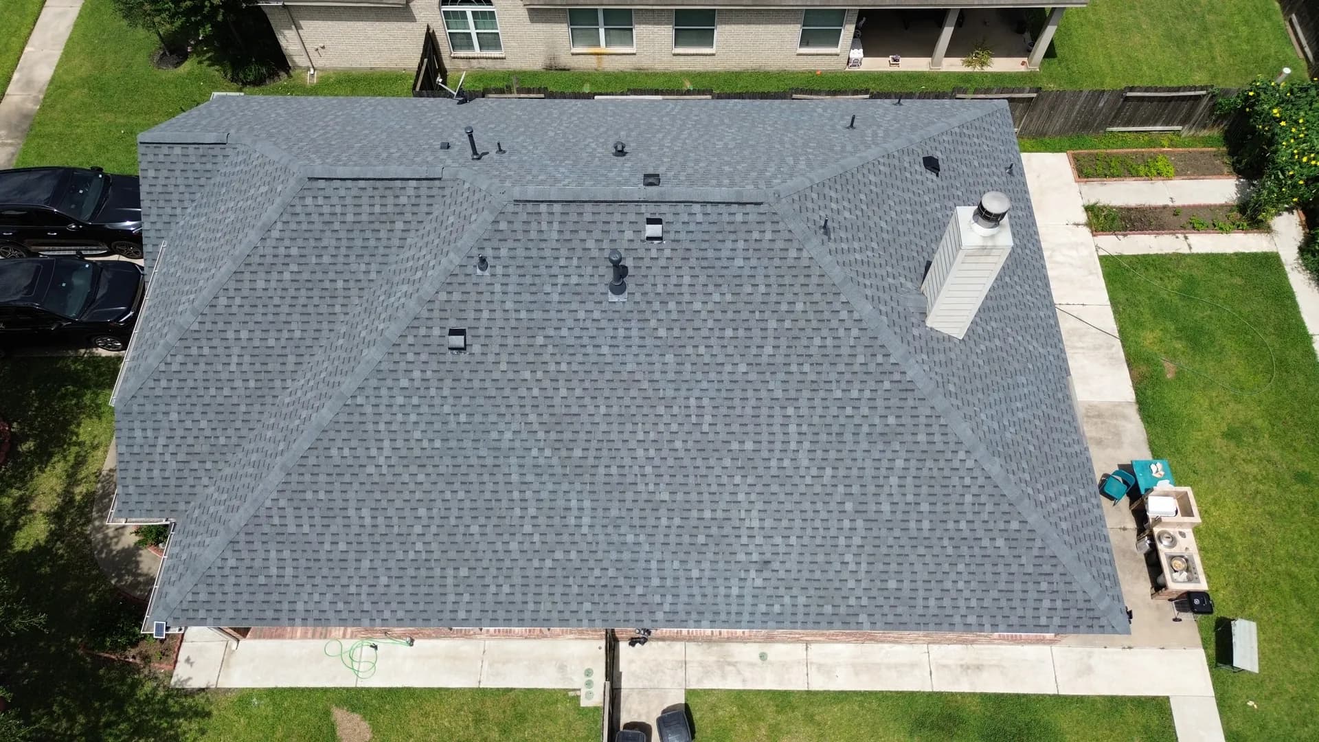 Aerial drone view of a completed gray shingle roof with a large square attic vent and cars parked in the driveway below