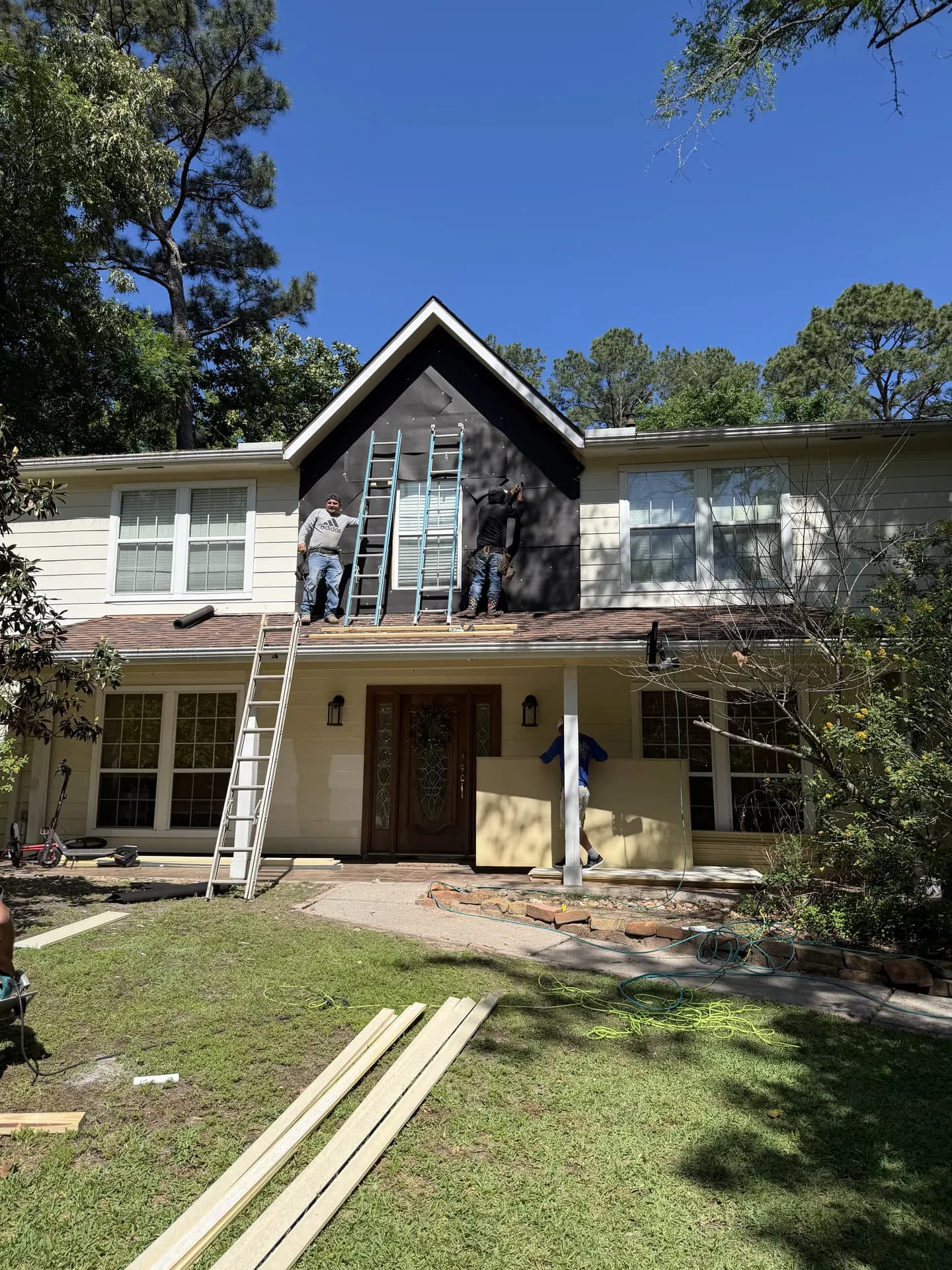 Full front view of a home in The Woodlands, TX mid-siding-replacement, with a ROYH Build Co crew working the gable and a worker carrying lumber across the yard