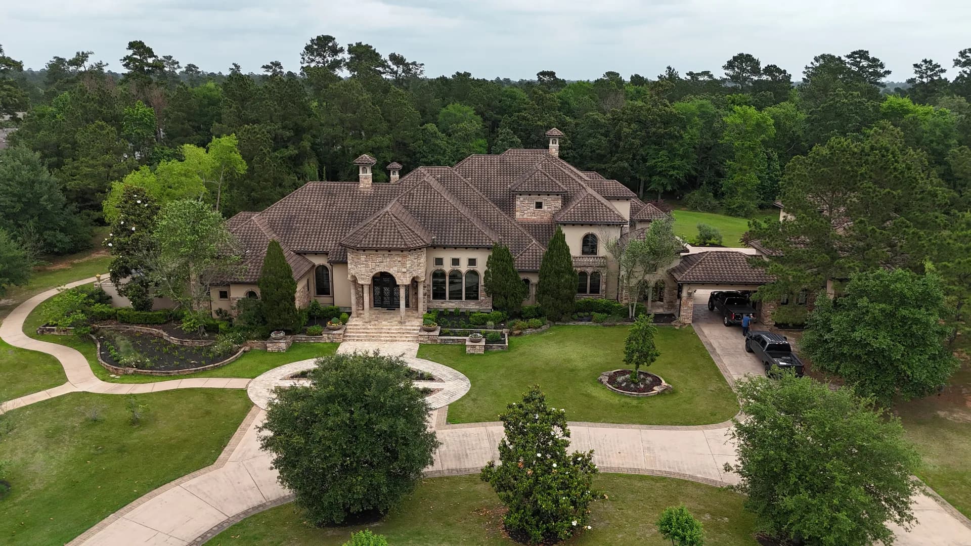 Aerial view of a large stucco and stone estate near The Woodlands, TX with a Mediterranean-style clay tile roof and a circular paver driveway